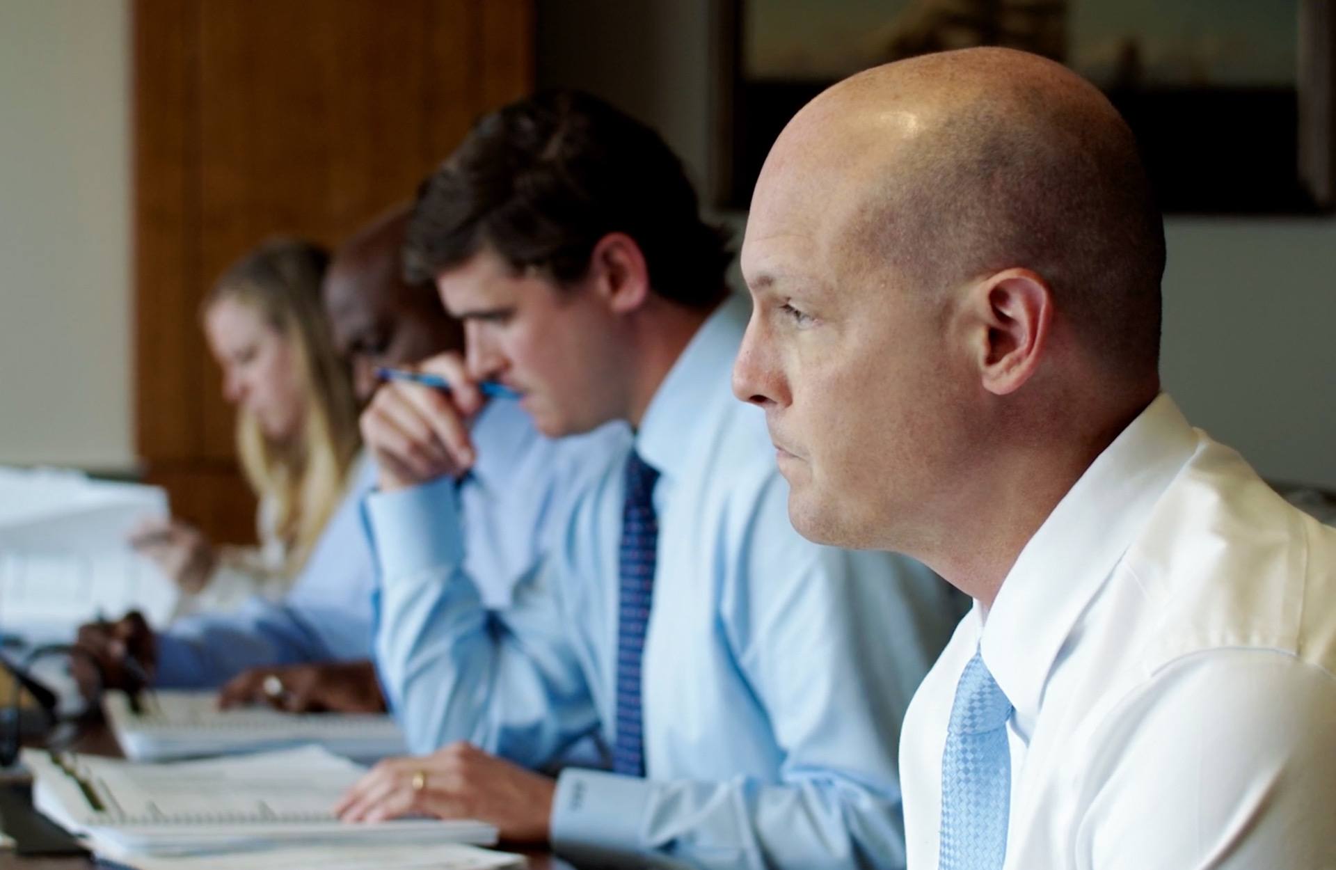 A BCP employee focused on the speaker during a meeting in the conference room while other employees review documents.